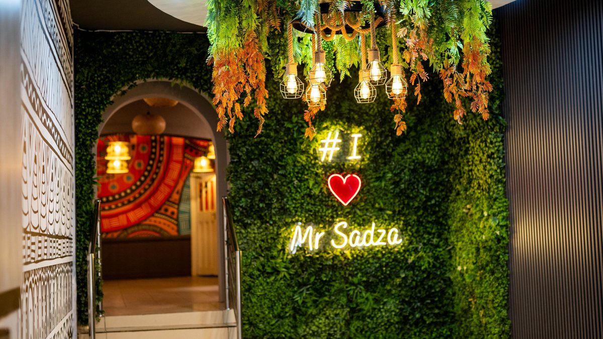 Mr Sadza illuminated sign on a green feature wall beneath hanging foliage and pendant lights.