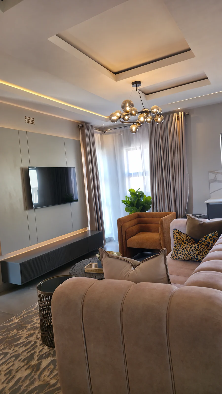 Living room with a sculpted ceiling, globe pendant lighting, wall-mounted television, upholstered seating and full-length curtains.